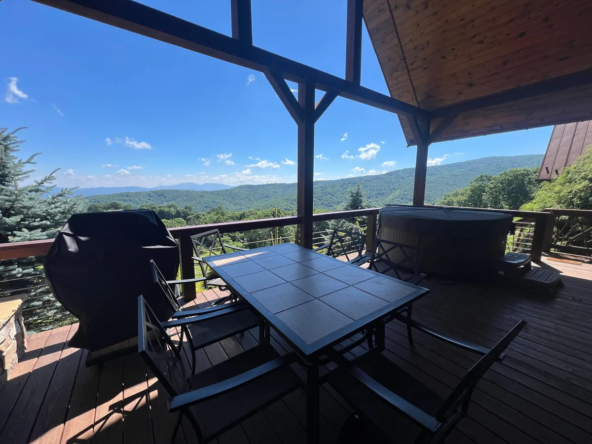 A spacious wooden deck featuring a dining table set for a group, a hot tub, and panoramic mountain views. The scene conveys a tranquil, social atmosphere perfect for gatherings, celebrations, and enjoying outdoor events. The deck's vantage point suggests a great spot for watching sunsets or fireworks displays.