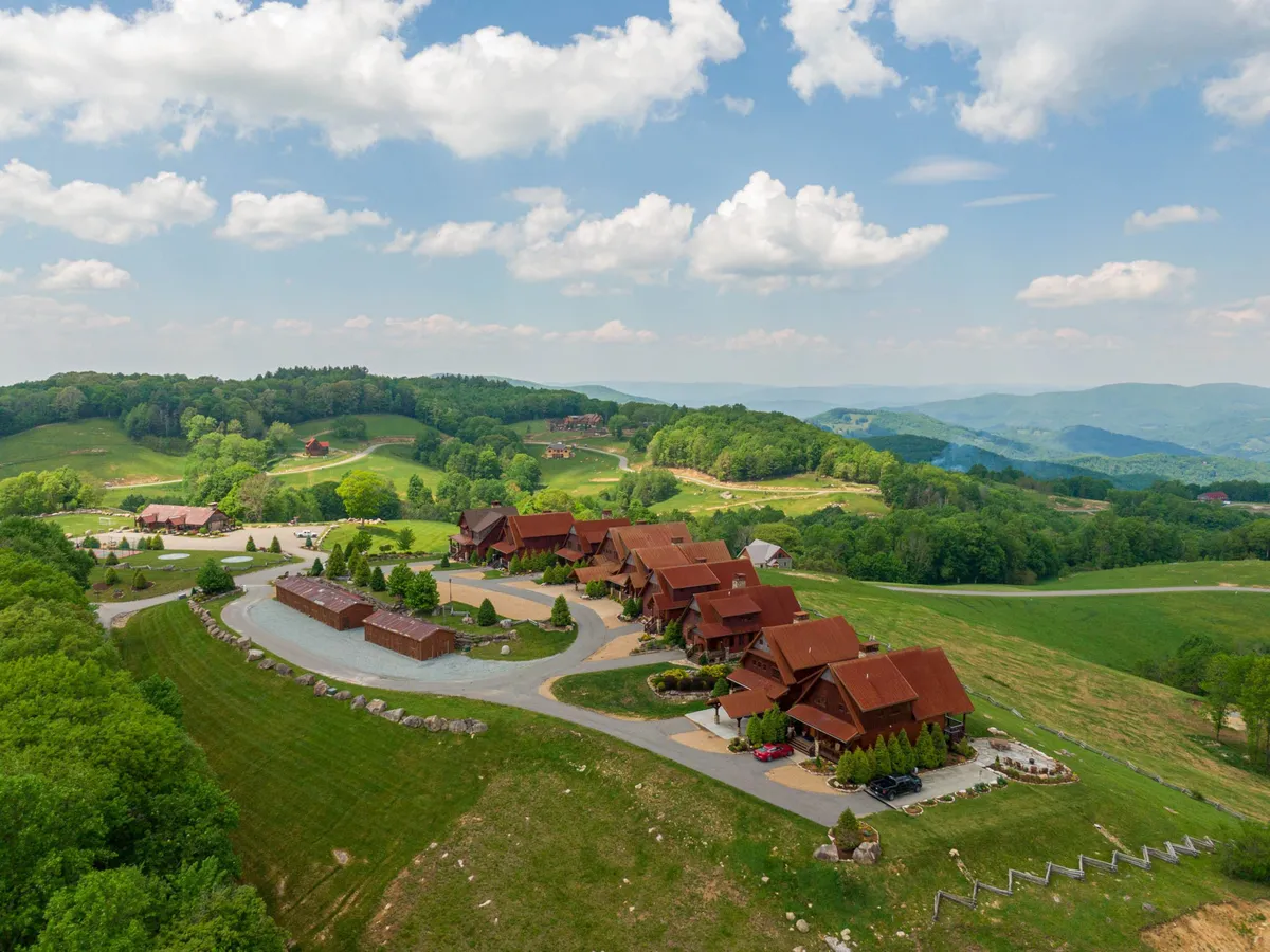 A scenic overview displays several wooden cabins with red roofs nestled among rolling green hills, landscaped gardens, and a winding road. The expansive outdoors invites exploration and communal activities.