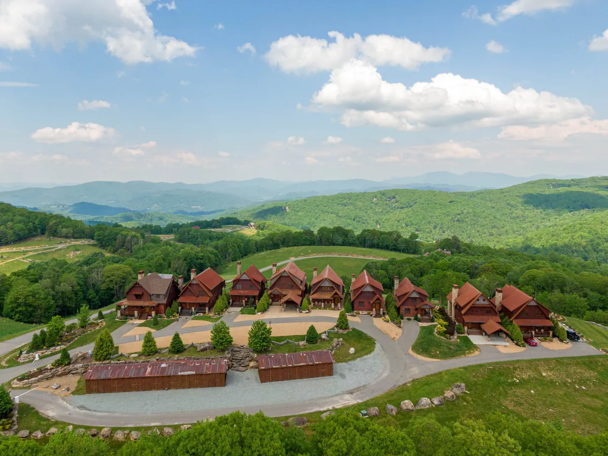 This exterior shot features a row of charming wooden cabins set amid a verdant, mountainous landscape with a clear blue sky and gravel driveway. The inviting atmosphere suggests communal outdoor activities and relaxation in nature.