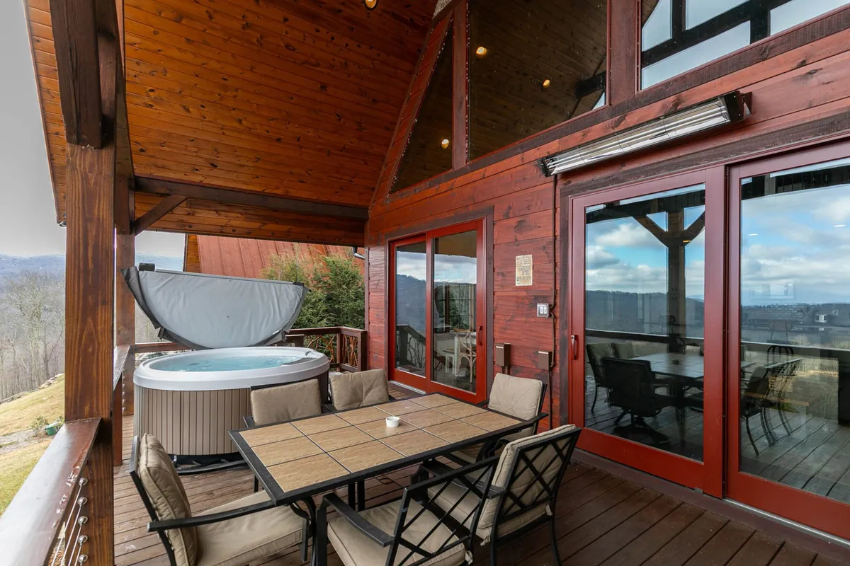 An exterior shot of a deck showcasing a hot tub and a dining table, all set against a beautiful mountain vista. The arrangement suggests an inviting space for outdoor dining, relaxing, or social gatherings, capturing the communal spirit of the property and surrounding area.