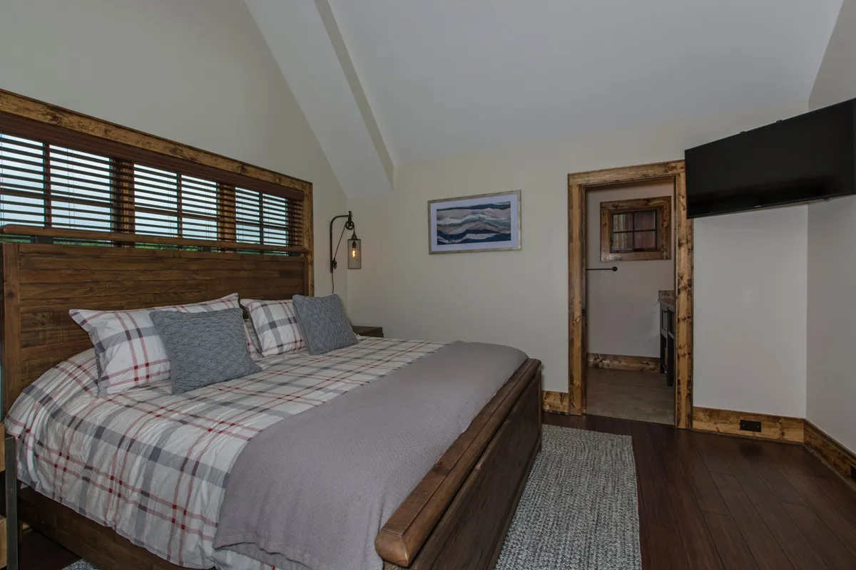 An inviting bedroom with a wooden bed frame, plaid bedding, wall artwork, and a hanging light fixture, illuminated by soft morning light. The ambiance is warm and welcoming, suitable for a master suite.