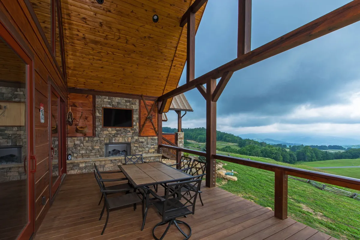 A spacious wooden deck featuring a stone outdoor fireplace, dining table, and sweeping mountain views. The arrangement highlights cozy gathering spots for both day and evening enjoyment, with the sunset backdrop enhancing the atmosphere.