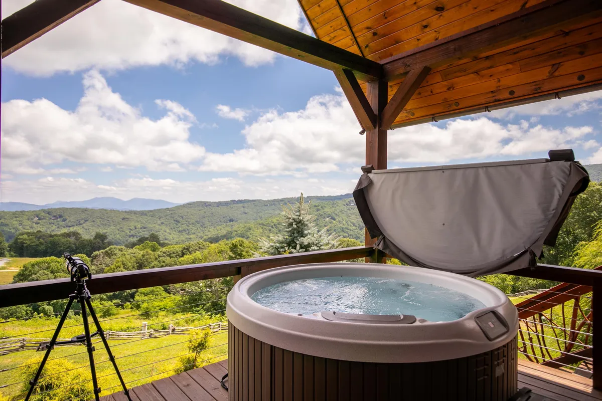 A tranquil scene showing a wooden deck with a hot tub facing the mountains, inviting guests to unwind while soaking in the view. The setting transitions beautifully from day to night, perfect for stargazing or morning coffee.