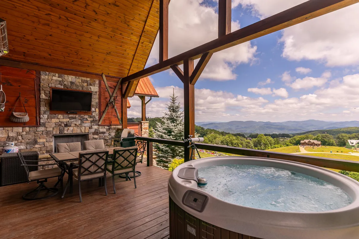 This image presents a luxurious deck setup featuring a hot tub, outdoor seating area, stone fireplace, wall-mounted TV, and breathtaking mountain views. The layout is inviting, with ample space for relaxing in the hot tub, enjoying a meal, or gathering around the fireplace.