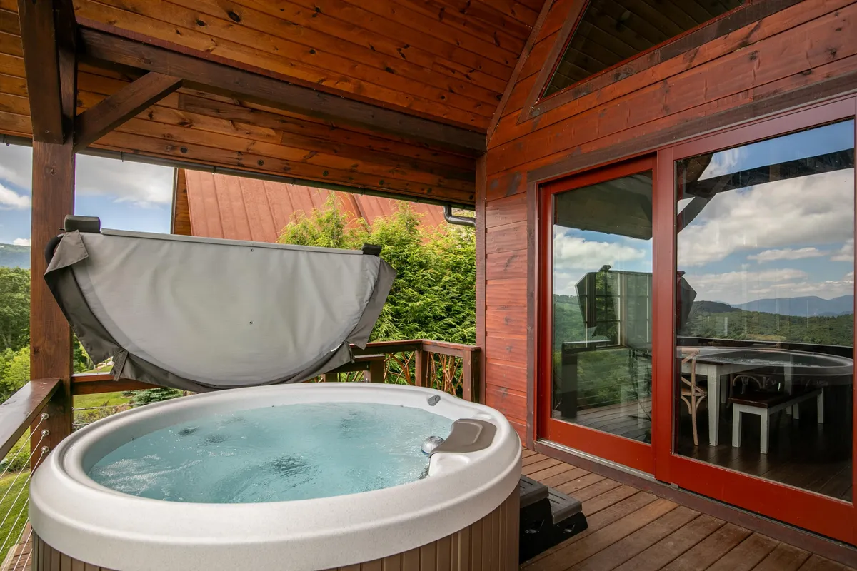 This image showcases the exterior wooden deck of the property, featuring a hot tub, large windows offering scenic views of the surrounding natural landscape, and a table for outdoor dining. The serene setting suggests relaxation and enjoyment of the outdoors in multiple seasons.