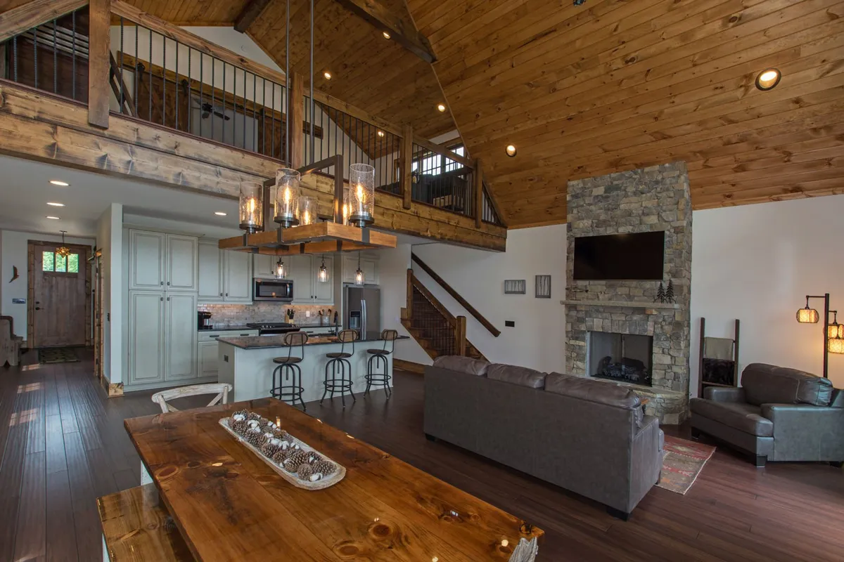 This image highlights the wooden dining table, a cozy stone fireplace, an open staircase, and part of the kitchen island. The layout underscores the fluid transition between dining, kitchen, and lounging spaces, while the glowing fireplace and ample seating convey warmth and sociability.
